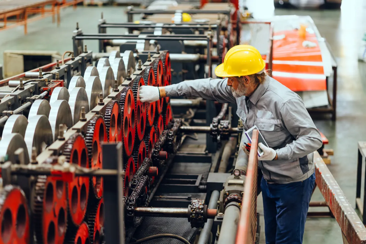 A worker in a hard hat inspects industrial machinery while holding a tablet.