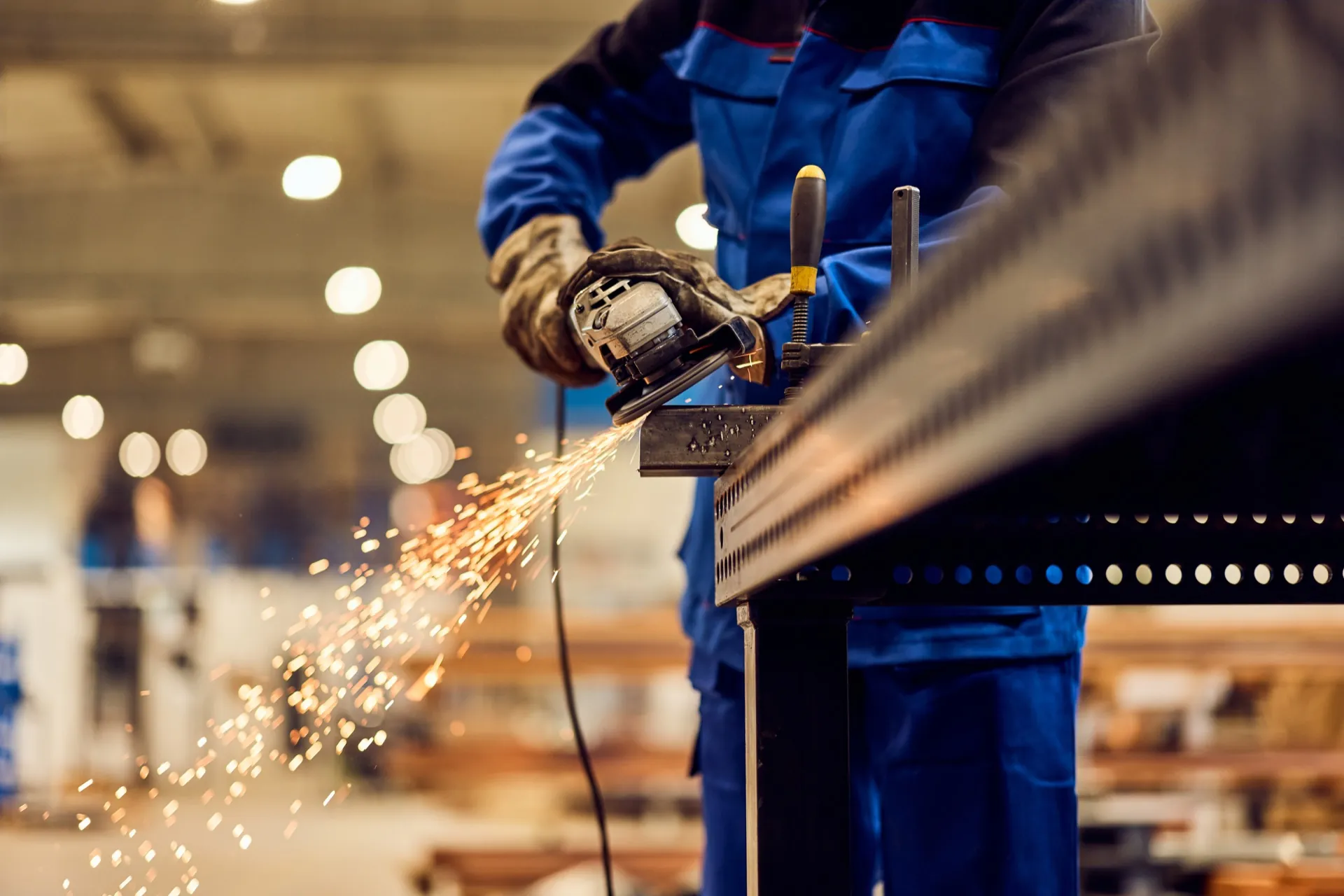 A worker uses an angle grinder on a clamped metal piece, sending sparks across a workshop.