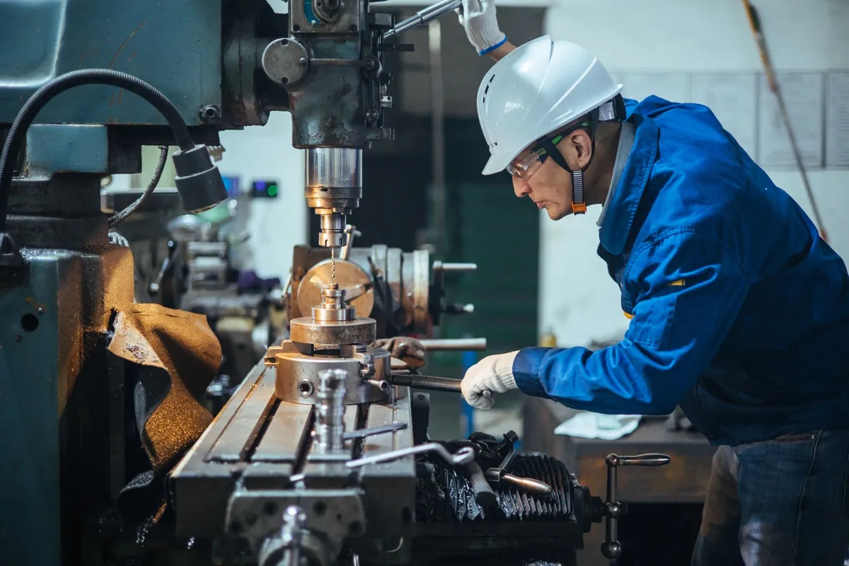 A worker wearing a hard hat and safety glasses operates a milling machine.
